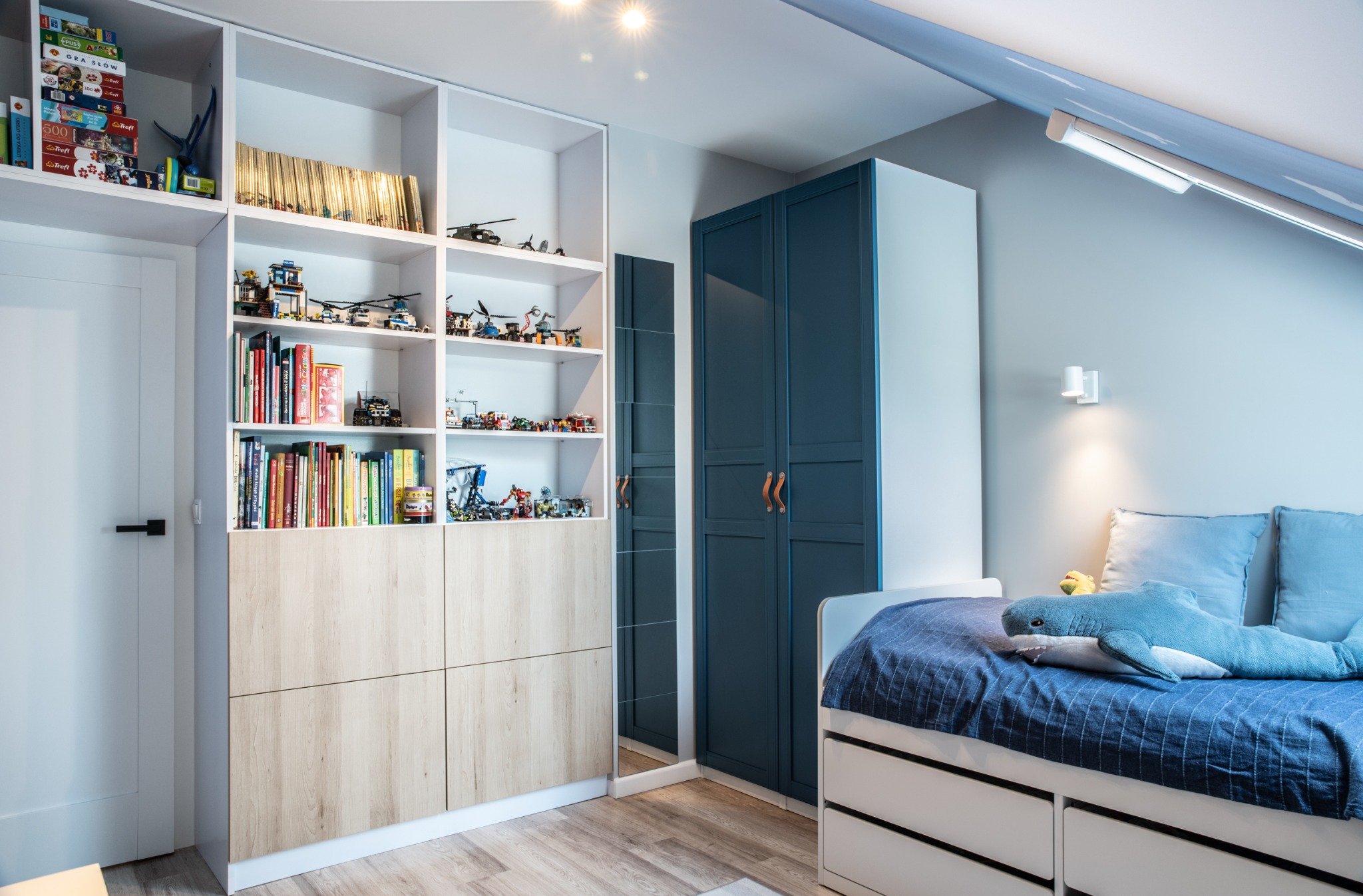 Pokój dziecięcy na poddaszu z białymi regałami wypełnionymi książkami i zabawkami Lego, granatową szafą z skórzanymi uchwytami, lustrem i łóżkiem z pluszowym rekinem.