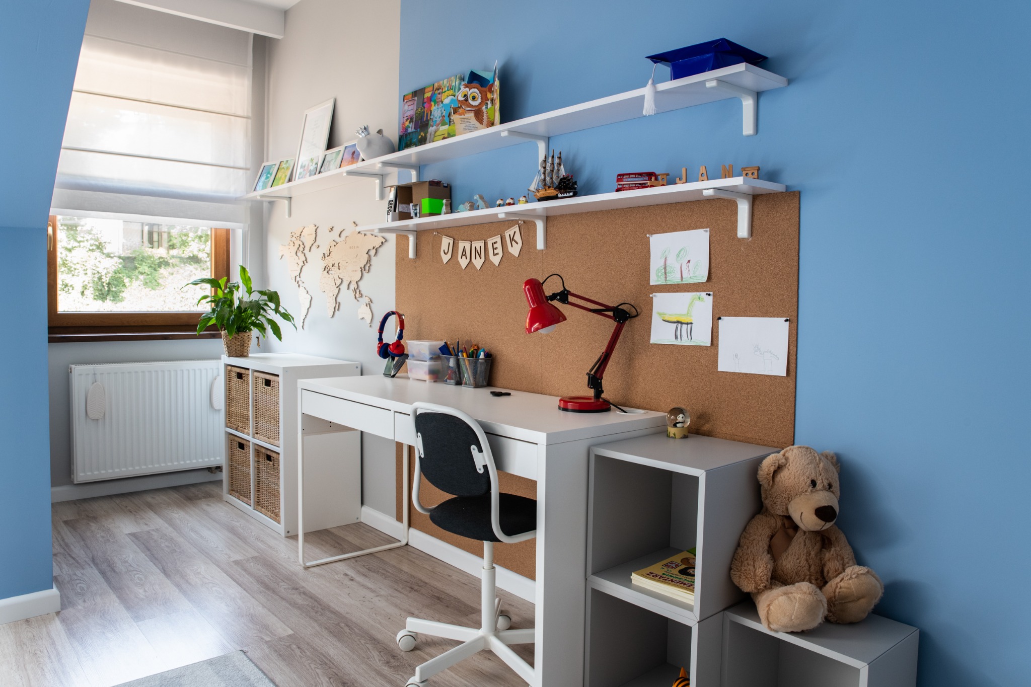 Pokój dziecięcy z białym biurkiem, krzesłem na kółkach, korkową tablicą z rysunkami, półkami z dekoracjami, pluszowym misiem i ścianą pomalowaną na dwa kolory: biały i niebieski.