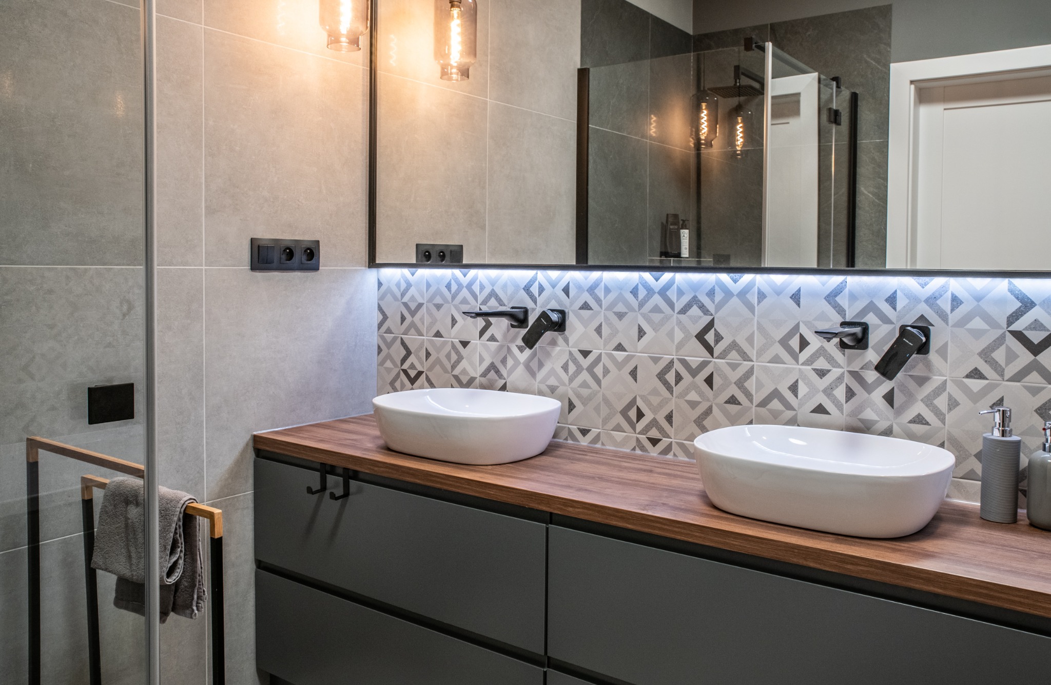 Nowoczesna łazienka z dwoma owalnymi umywalkami nablatowymi, czarnymi bateriami ściennymi, drewnianym blatem i szafką w kolorze grafitowym, podświetlane lustro oraz geometryczne płytki na ścianie.