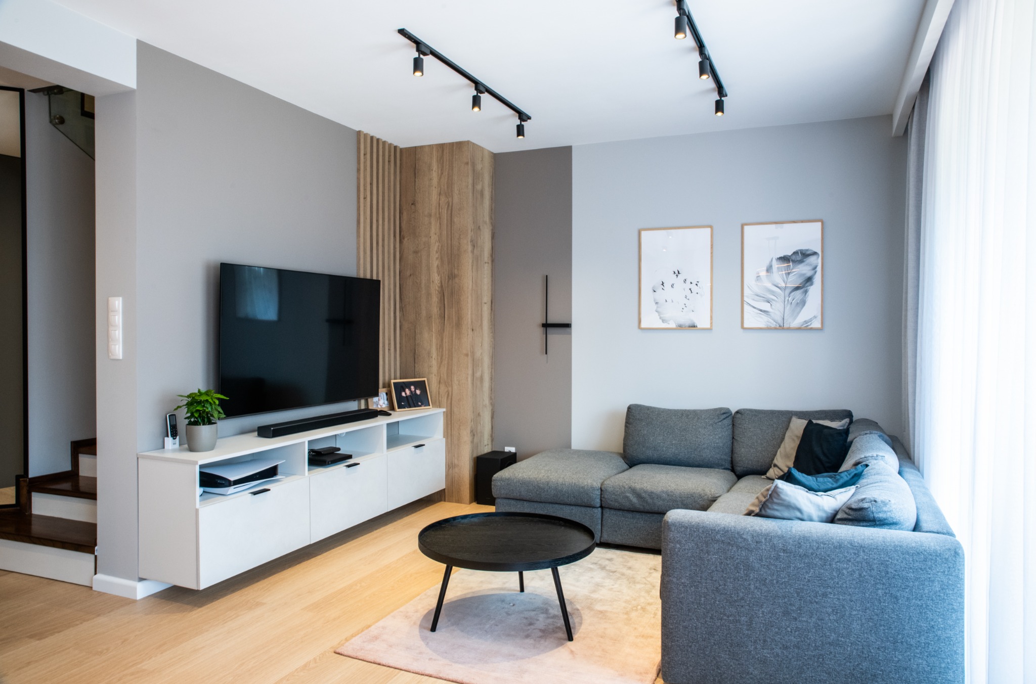 Nowoczesny salon z szarą narożną sofą, okrągłym czarnym stolikiem kawowym, białą szafką RTV, drewnianą ścianką dekoracyjną i oświetleniem szynowym.