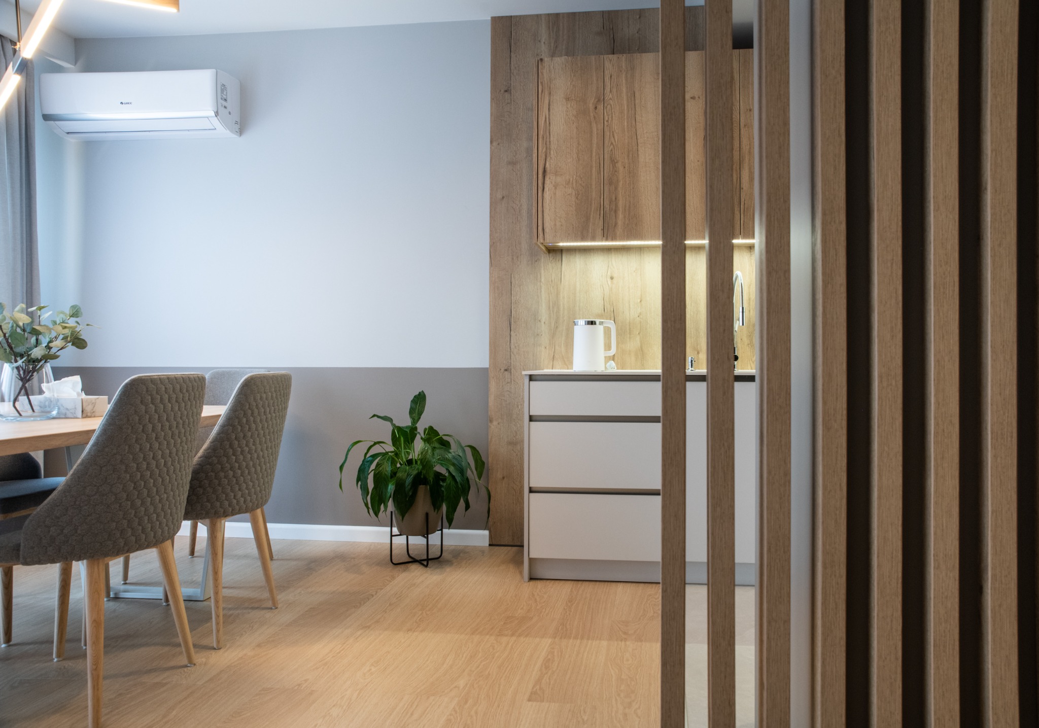 Nowoczesne wnętrze: widok na jadalnię z szarymi krzesłami i drewnianym stołem, oraz kuchnię z białymi szafkami i drewnianymi akcentami, oddzieloną drewnianymi lamelami. Na ścianie klimatyzator.
