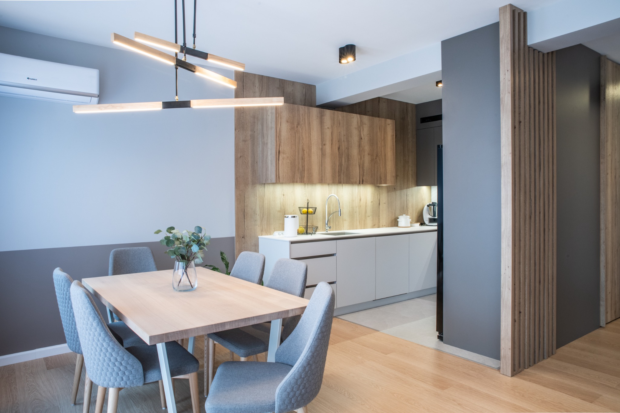 Nowoczesna jadalnia z drewnianym stołem otoczonym sześcioma szarymi krzesłami, minimalistyczna lampa sufitowa, kuchnia w tle z drewnianymi i szarymi szafkami, klimatyzator na ścianie.