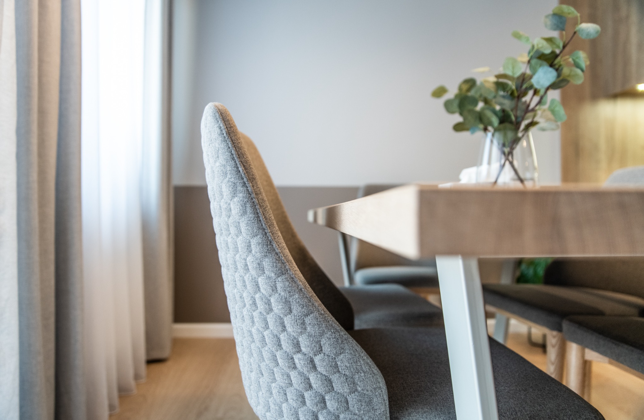 Szary, pikowany fotel przy drewnianym stole z bukietem eukaliptusa w szklanym wazonie, nowoczesne wnętrze jadalni z jasnym oknem.