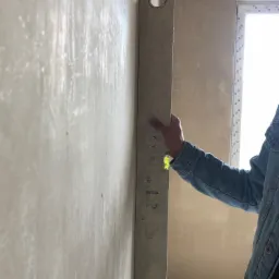 Świeżo nałożony, jasny tynk na ścianie, sprawdzany pionową poziomicą przez osobę w dżinsowej kurtce, widoczne okno w tle.