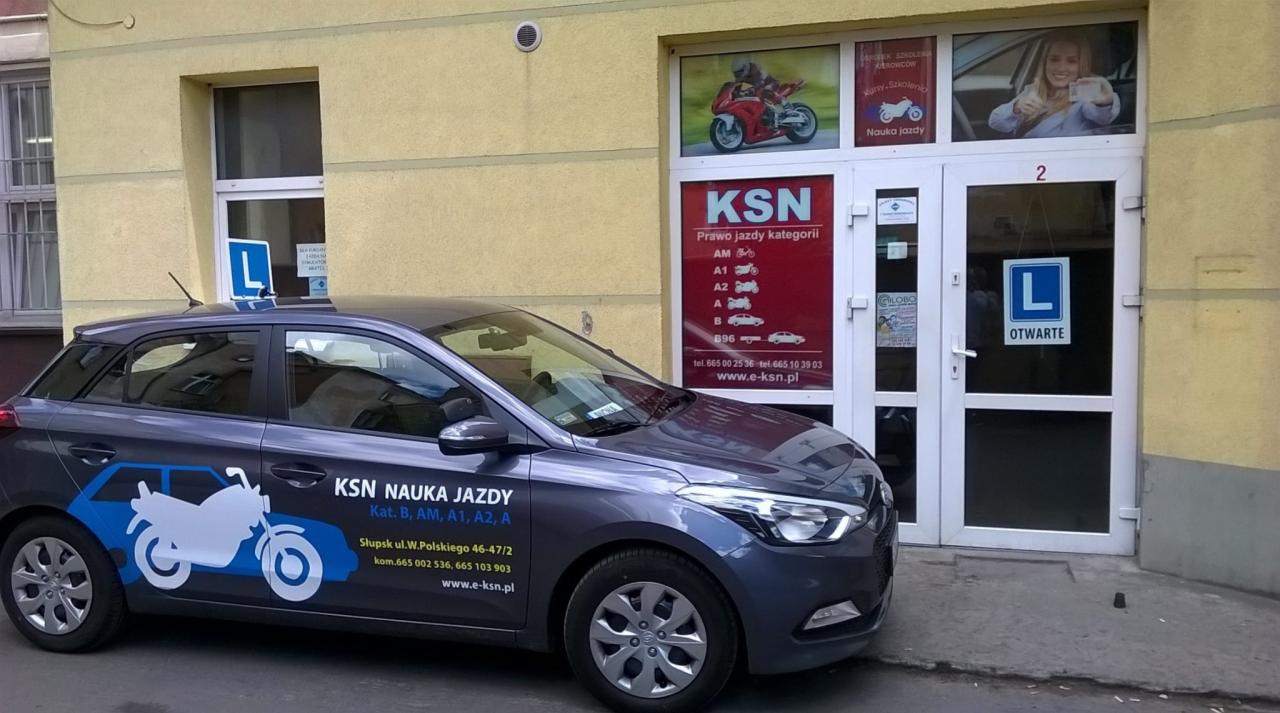 Samochód z naklejkami motywu szkoły jazdy KSN przed wejściem do biura, w tle szyld z informacjami o kategoriach prawa jazdy.