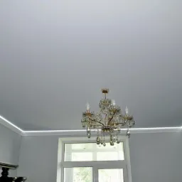 Gładki, jasnoszary sufit z subtelnym oświetleniem LED wzdłuż krawędzi, ozdobny żyrandol kryształowy, ściany w dwóch odcieniach szarości z wzorzystą tapetą i oknem.