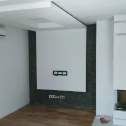 Nowoczesne wnętrze po remoncie z białymi ścianami, drewnianą podłogą, grzejnikiem panelowym, klimatyzatorem, obudową kominka i dekoracyjnym panelem ściennym z kamienia.