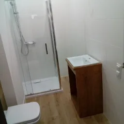 Nowoczesna łazienka z kabiną prysznicową ze srebrnymi ramami, drewnianą szafką pod umywalką i toaletą, wykończona białymi płytkami i drewnopodobną podłogą.