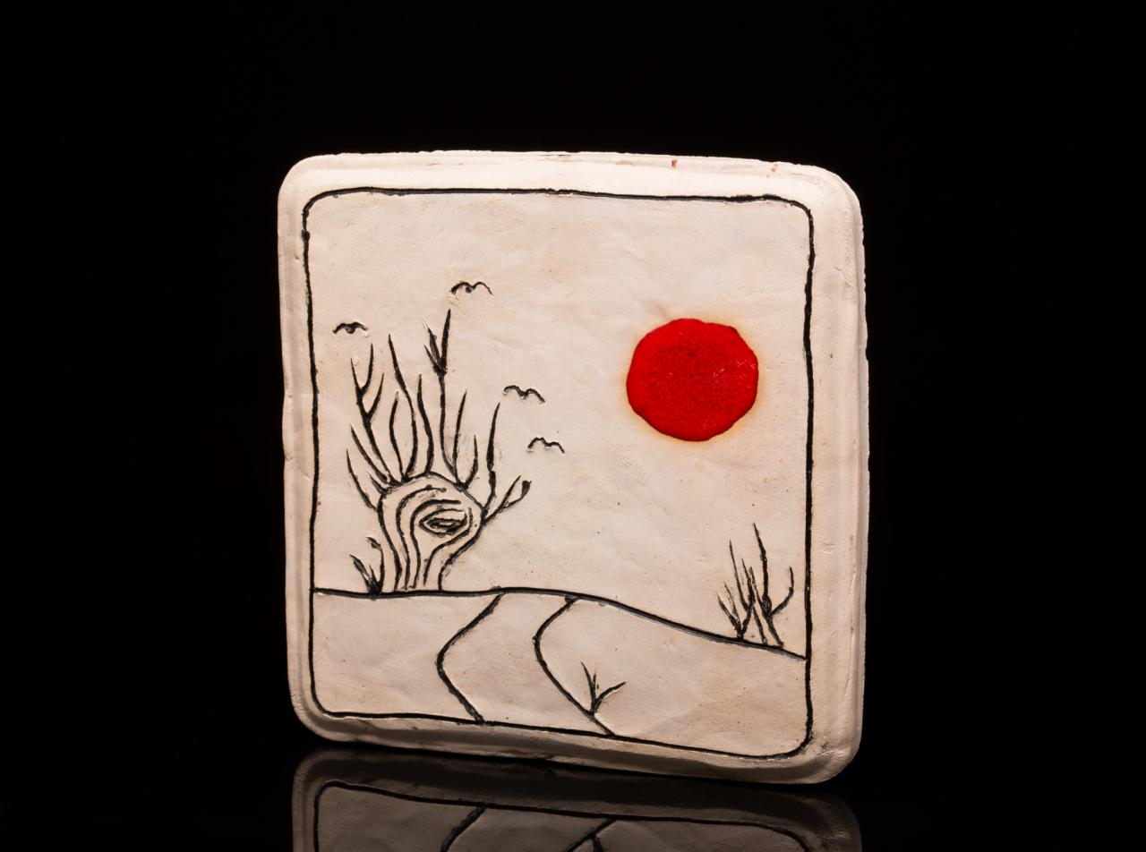 Dekoracyjny, ręcznie wykonany ceramiczny kafelek z wypukłym czerwonym słońcem i czarnym rysunkiem drzewa oraz drogi na białym tle, na czarnym, błyszczącym podłożu.