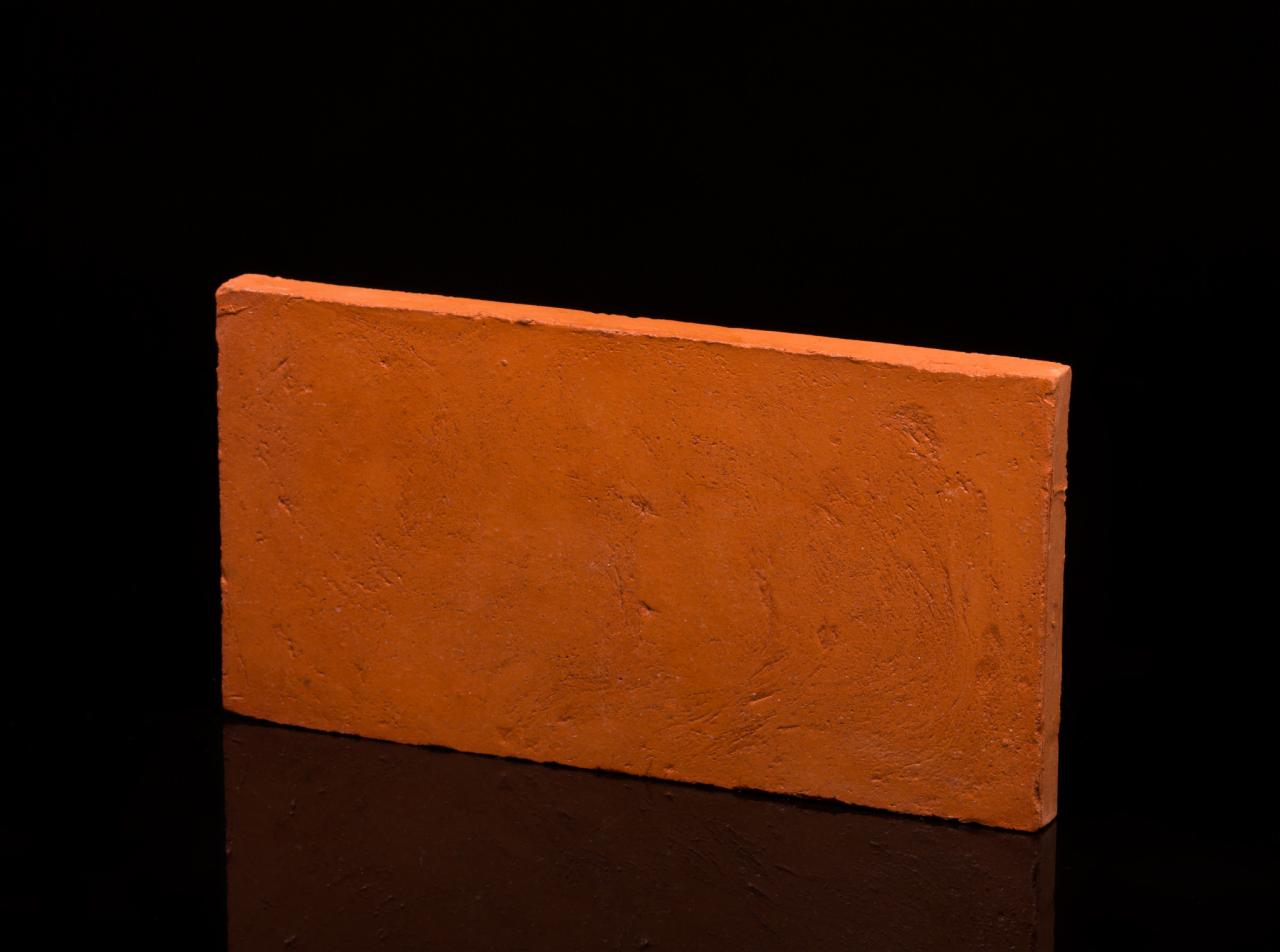 Pojedyncza, terakotowa płytka podłogowa z widoczną, lekko chropowatą fakturą, ułożona na błyszczącej czarnej powierzchni, odbijająca światło