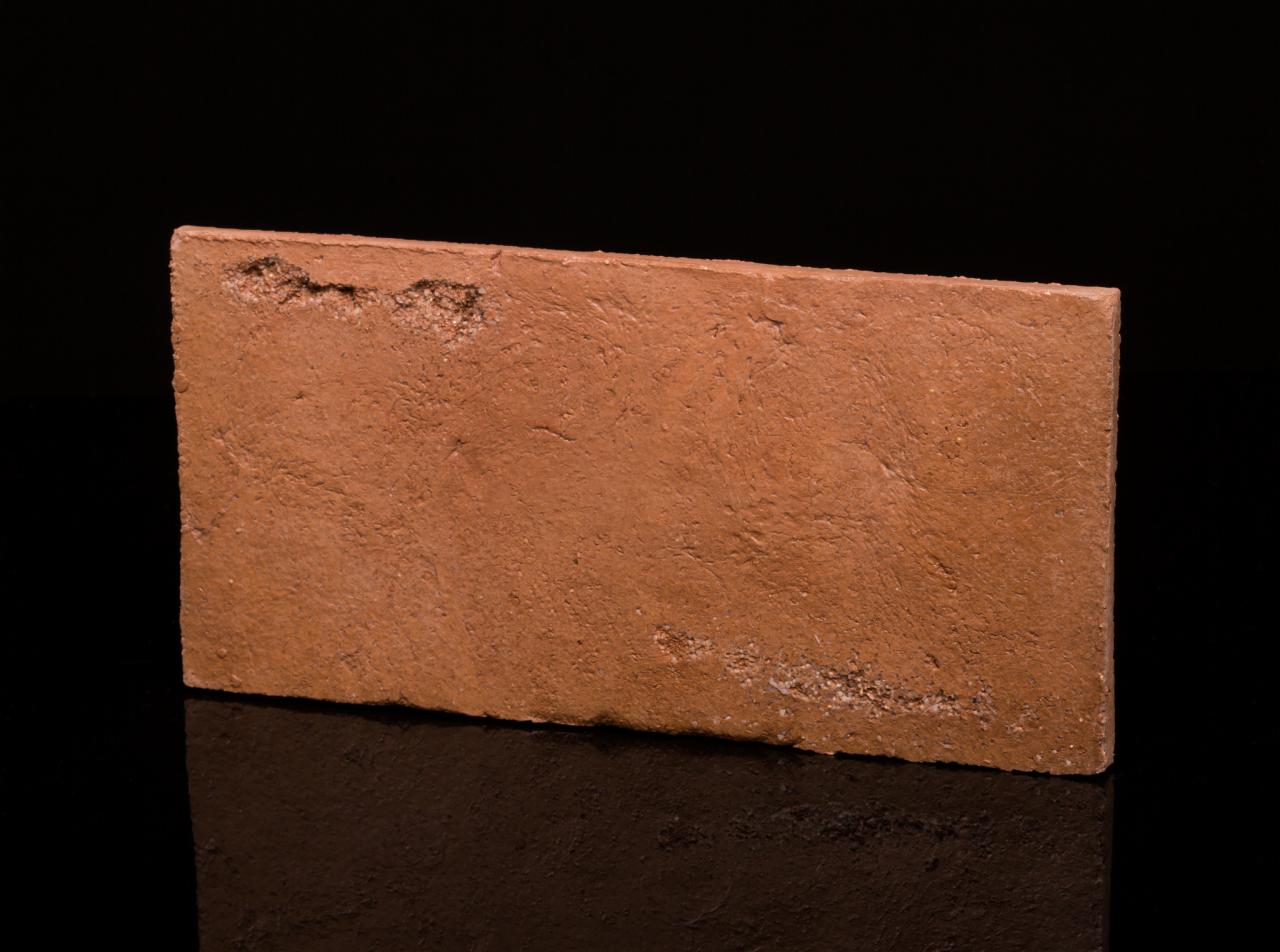 Pojedyncza, terakotowa płytka podłogowa z widoczną, nieregularną fakturą i drobnymi ubytkami na krawędzi, leżąca na czarnym, błyszczącym tle.