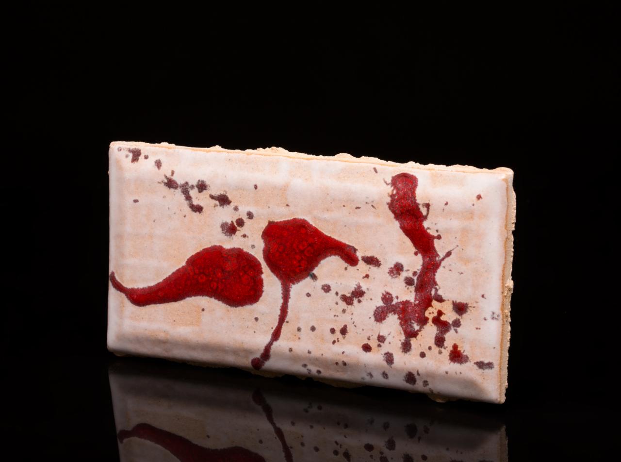 Ceramiczna płytka podłogowa z abstrakcyjnym wzorem czerwonych plam na beżowym tle, prezentowana na czarnym, błyszczącym podłożu.