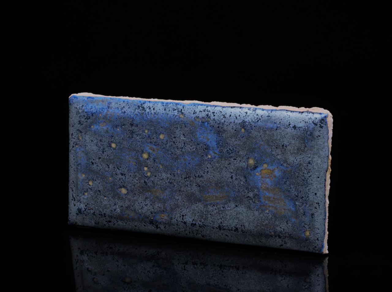 Niebiesko-szara próbka panela z nieregularną fakturą i drobnymi, złotymi plamkami, umieszczona na czarnym, błyszczącym tle.