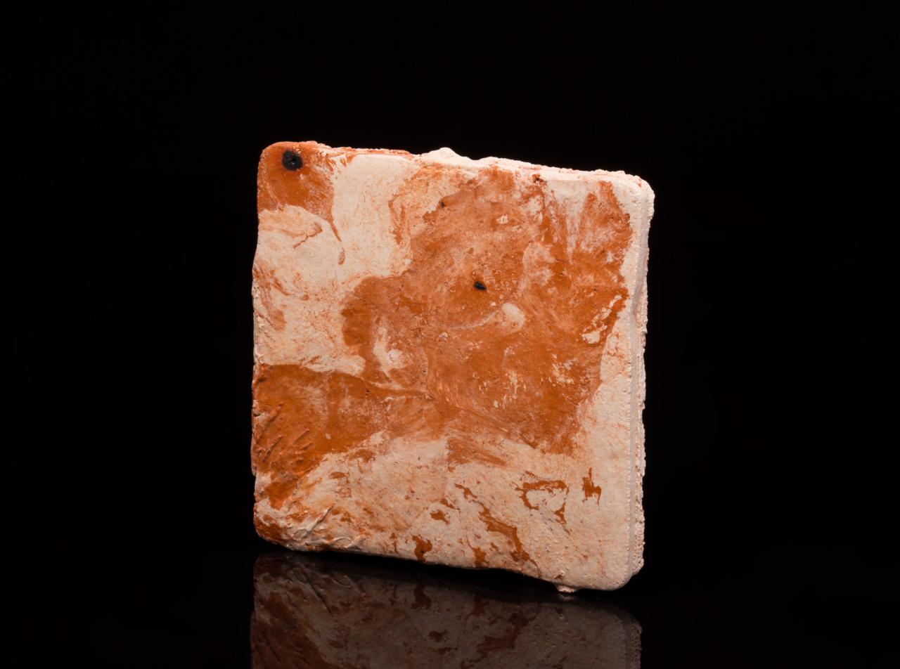 Kwadratowa płytka terakotowa z nieregularną, marmurkową fakturą w odcieniach pomarańczy i bieli, widoczna na czarnym, błyszczącym tle.
