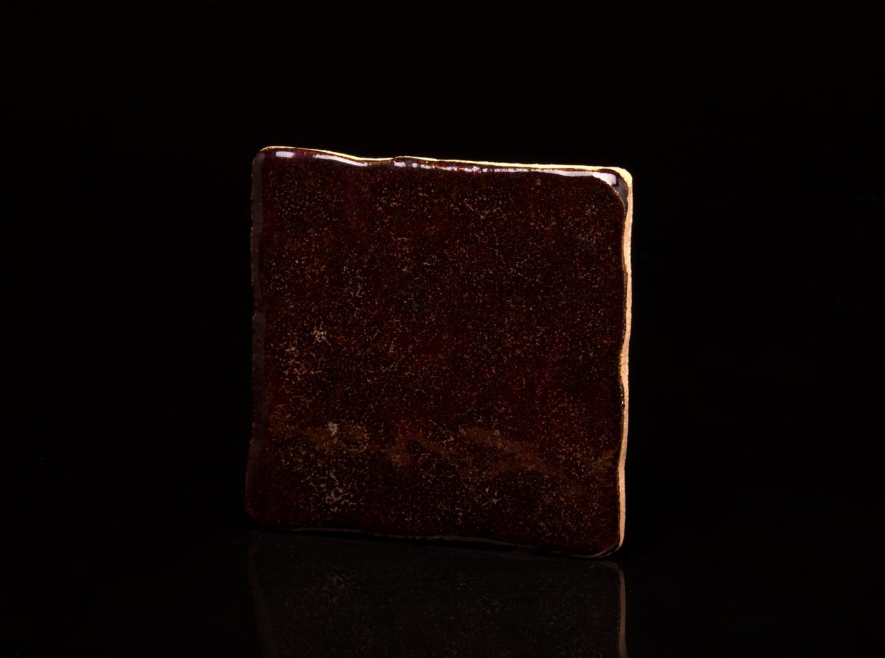 Kwadratowa płytka w kolorze ciemnego brązu z nieregularnymi krawędziami i połyskującą powierzchnią, prezentowana na czarnym tle z odbiciem.