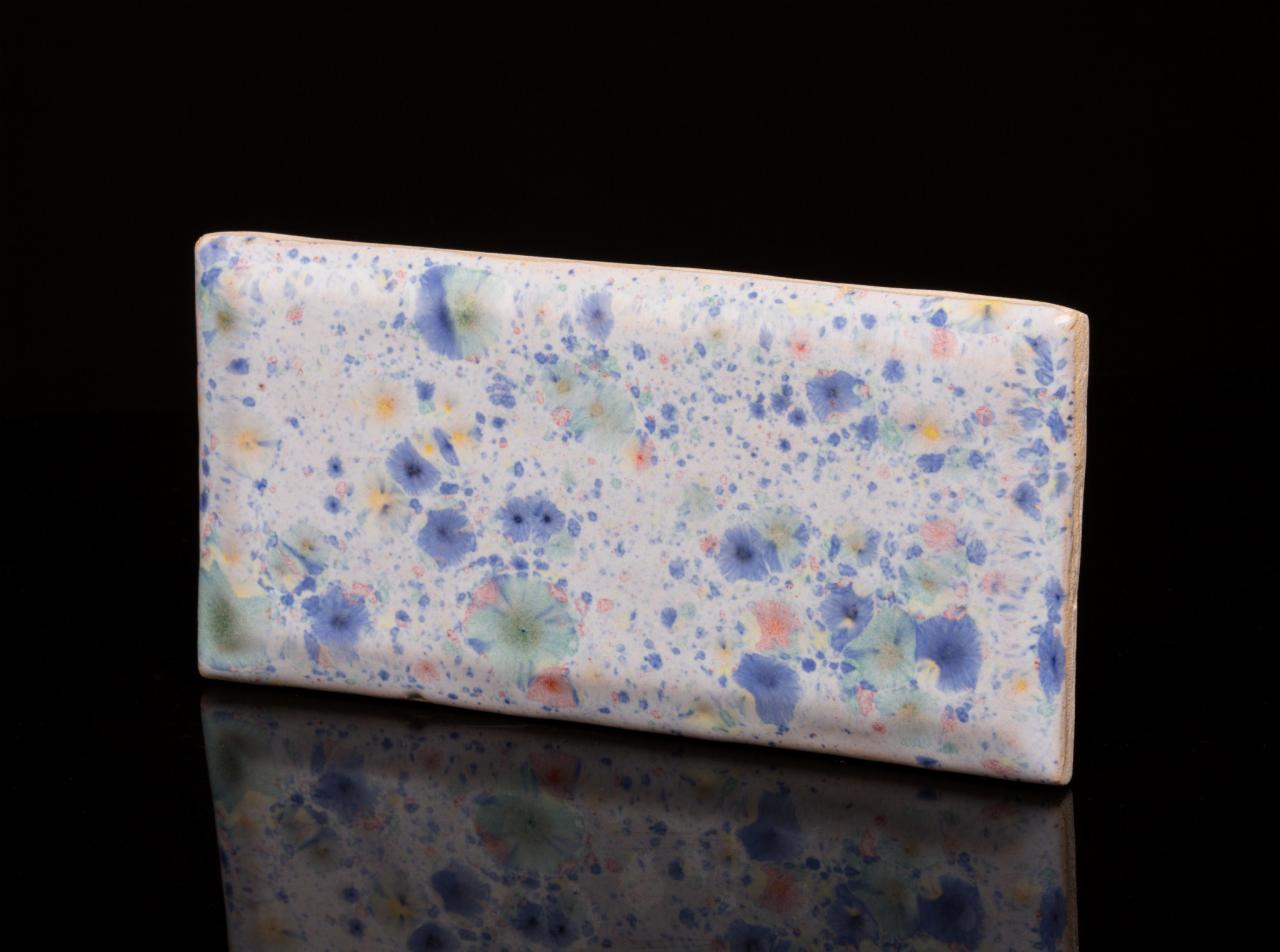 Ceramiczna płytka z abstrakcyjnym, kwiatowym wzorem w odcieniach błękitu, zieleni i żółci na białym tle, leżąca na czarnym, błyszczącym podłożu.