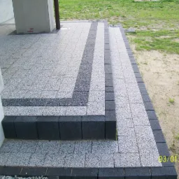 Schody wejściowe wyłożone szarą kostką brukową z ciemnoszarym obramowaniem i pasami, widoczne z boku budynku.