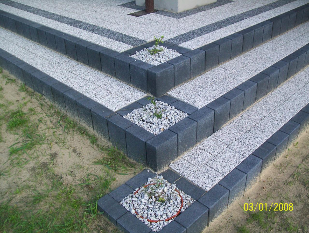 Schody zewnętrzne wyłożone jasną kostką z ciemnymi obrzeżami, z małymi rabatami wypełnionymi białymi kamykami i sadzonkami.