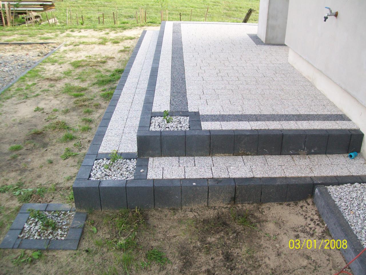 Schody wejściowe wyłożone kostką brukową w odcieniach szarości, z elementami dekoracyjnymi wypełnionymi białym grysem i młodymi roślinami, widoczne na tle trawnika i fragmentu elewacji budynku.