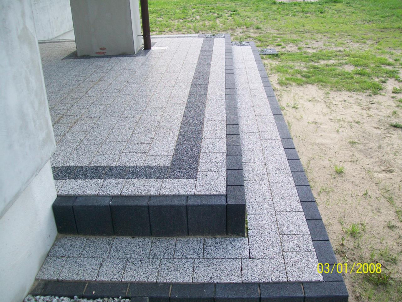 Schody wejściowe wyłożone szarą kostką brukową z ciemnoszarym obramowaniem i pasami, widoczne z boku budynku.