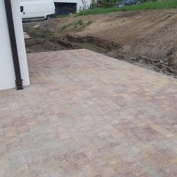 PROBUD Krzysztof Gołda - Nowo ułożona, wielobarwna kostka brukowa w odcieniach beżu i czerwieni przylegająca do ściany budynku z rurą spustową, z widocznym fragmentem terenu przygotowanego pod dalsze prace ziemne.