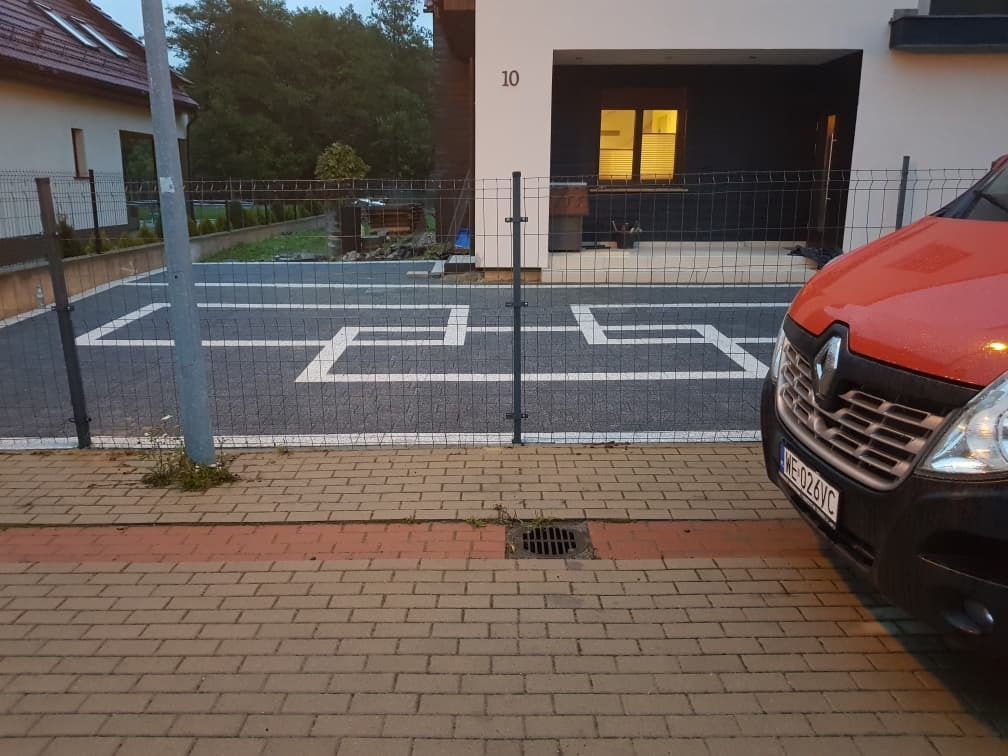 Nowo wyłożony, ciemnoszary parking z kostki brukowej Semmelrock z białymi liniami wyznaczającymi miejsca postojowe, widok zza metalowego ogrodzenia i częściowo zasłaniającego go czerwonego...