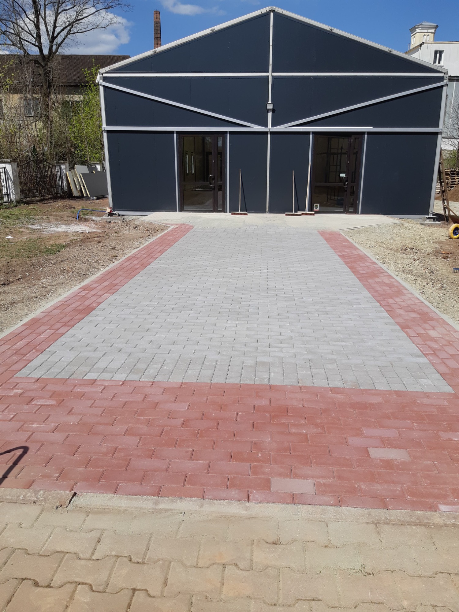 Ułożona kostka brukowa w kolorze szarym z obramowaniem z czerwonej kostki, prowadząca do nowoczesnego, szarego budynku z przeszklonymi drzwiami wejściowymi, widoczne dwie miotły oparty o budynek.