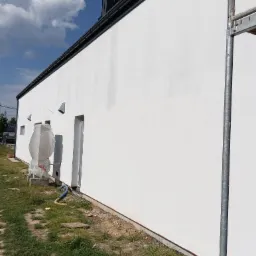 Boczny widok świeżo pomalowanej na biało elewacji budynku z widocznymi oknami i drzwiami zabezpieczonymi folią, z instalacją owiniętą folią ochronną i fragmentem rusztowania po prawej stronie.