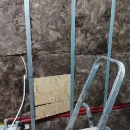 Ściana ocieplona wełną mineralną z widocznymi profilami metalowymi, fragmentem instalacji wodnej i drabiny malarskiej.