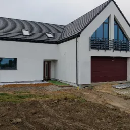 Nowoczesny dom jednorodzinny z białą elewacją, ciemną dachówką i bordową bramą garażową. Widoczne okna z ciemnymi ramami, mały balkon z czarną balustradą i fragment niezagospodarowanego terenu...