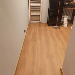 Nowo ułożone panele podłogowe w korytarzu, widoczne listwy przypodłogowe, okno z żaluzjami i fragment ściany.