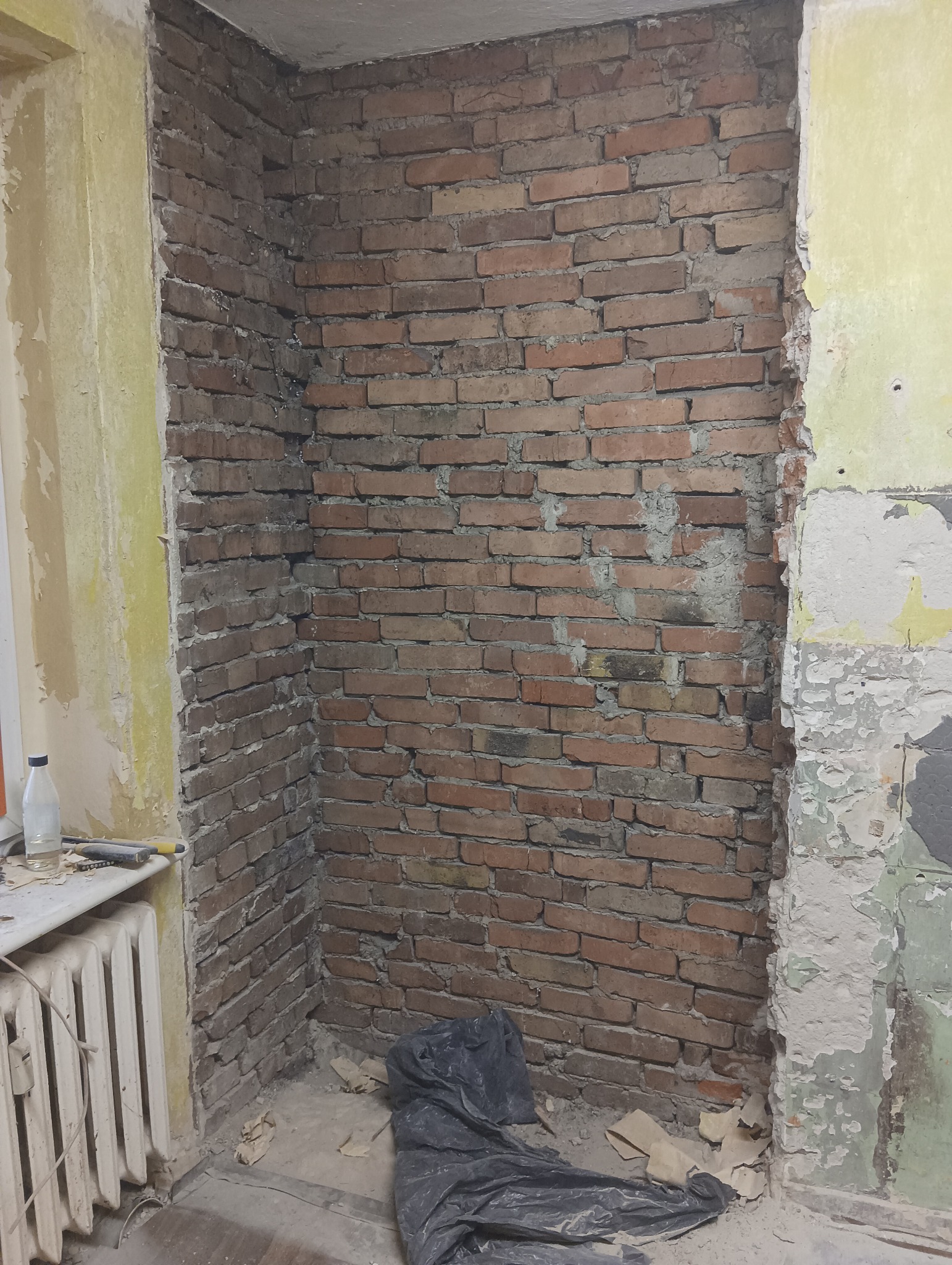 Odsłonięta ceglana ściana w trakcie remontu mieszkania, widoczne fragmenty starej farby na ścianach, grzejnik i butelka wody na parapecie, oraz folia ochronna na podłodze.