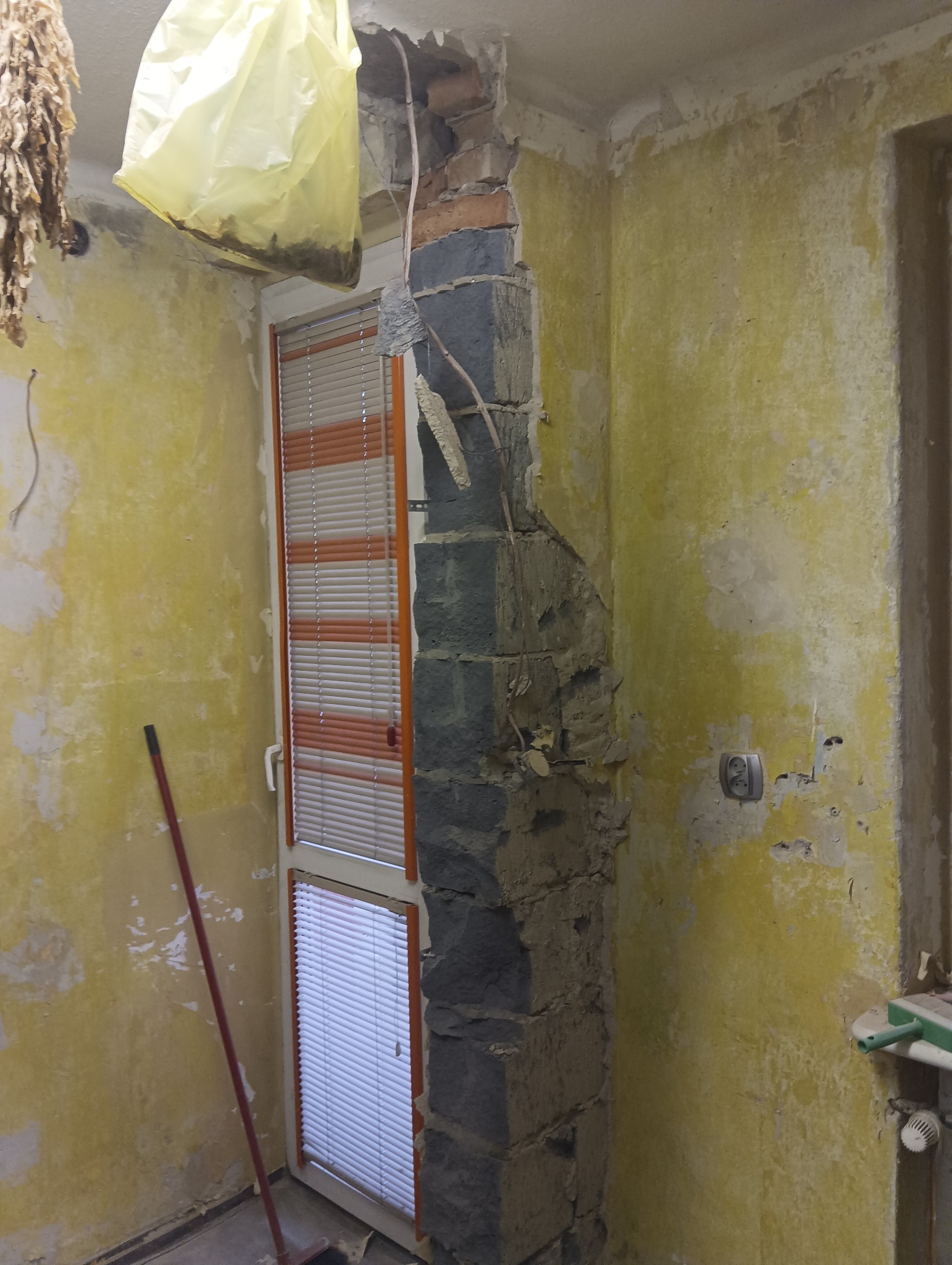 Demontaż ściany z cegły w trakcie remontu mieszkania, widoczne odsłonięte przewody elektryczne, resztki starej farby na ścianie i okno z żaluzjami.