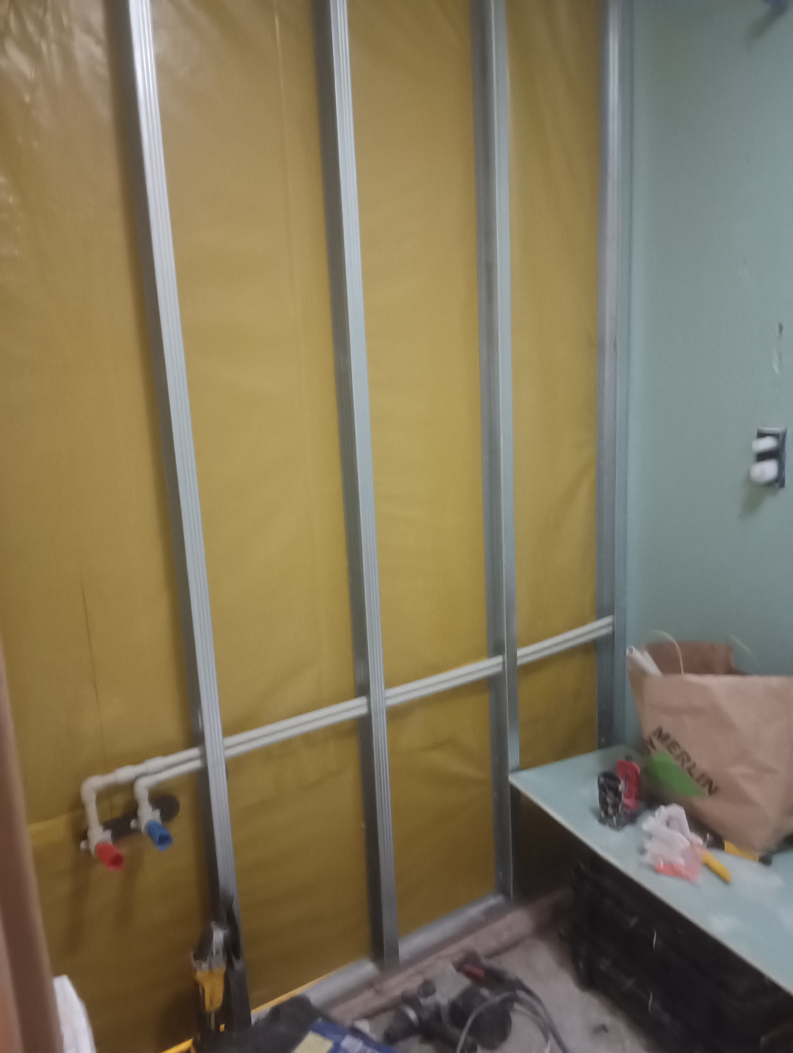 Wnętrze w trakcie remontu: metalowy stelaż pod zabudowę z płyt gipsowo-kartonowych na żółtej folii izolacyjnej, instalacja wodna z zaworami ciepłej i zimnej wody, narzędzia oraz torba budowlana...