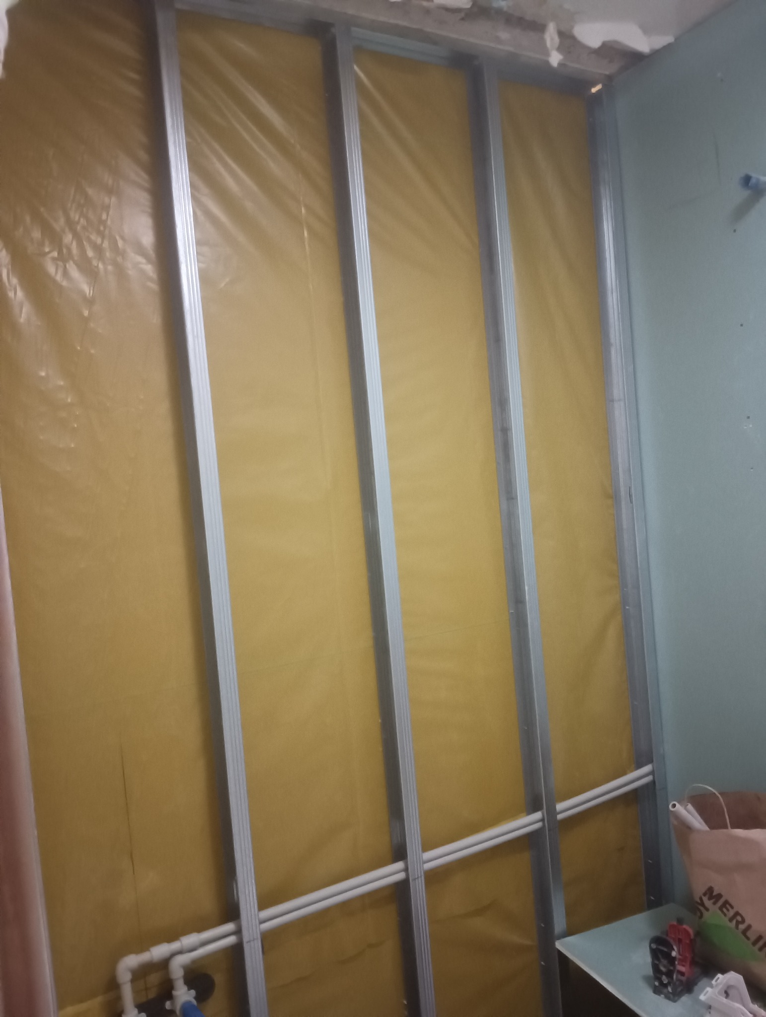 Wnętrze w trakcie remontu: metalowa konstrukcja szkieletowa na żółtej folii izolacyjnej, widoczne rury instalacyjne i torba z logo marketu budowlanego.