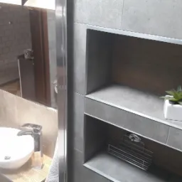 Nowoczesna łazienka z szarą glazurą, wnęką z drewnianym wykończeniem, małą rośliną w białej doniczce oraz lustrem odbijającym wnętrze z beżową cegłą.