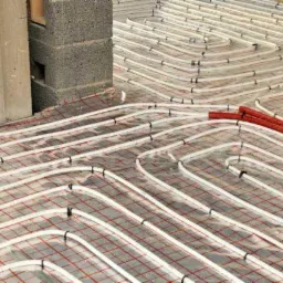 Rozłożone rury ogrzewania podłogowego na czerwonej siatce z widocznymi elementami mocującymi i fragmentami ścian w surowym stanie.