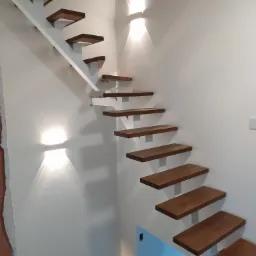 Nowoczesne, wiszące schody z drewnianymi stopniami i białym wspornikiem, oświetlone kinkietami na białej ścianie, widok z dołu.