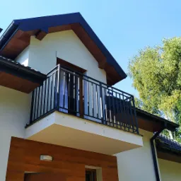 Nowoczesny balkon z czarną metalową balustradą na tle jasnej elewacji domu i błękitnego nieba.