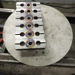 Rząd identycznych, metalowych uchwytów z widocznymi, kolorowymi śladami spawania, ułożonych na okrągłym metalowym stole warsztatowym.