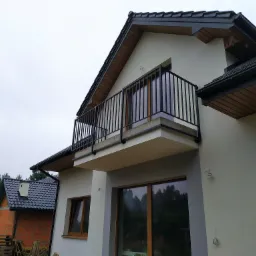 Nowoczesny dom z balkonem zabezpieczonym czarną, metalową konstrukcją, widok z dołu na tle pochmurnego nieba.