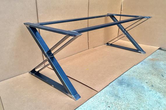 Stalowa konstrukcja stołu z nogami w kształcie litery Z, spawana z profili kwadratowych, surowy przemysłowy wygląd, widoczne spawy, tło z kartonu i betonowej podłogi.