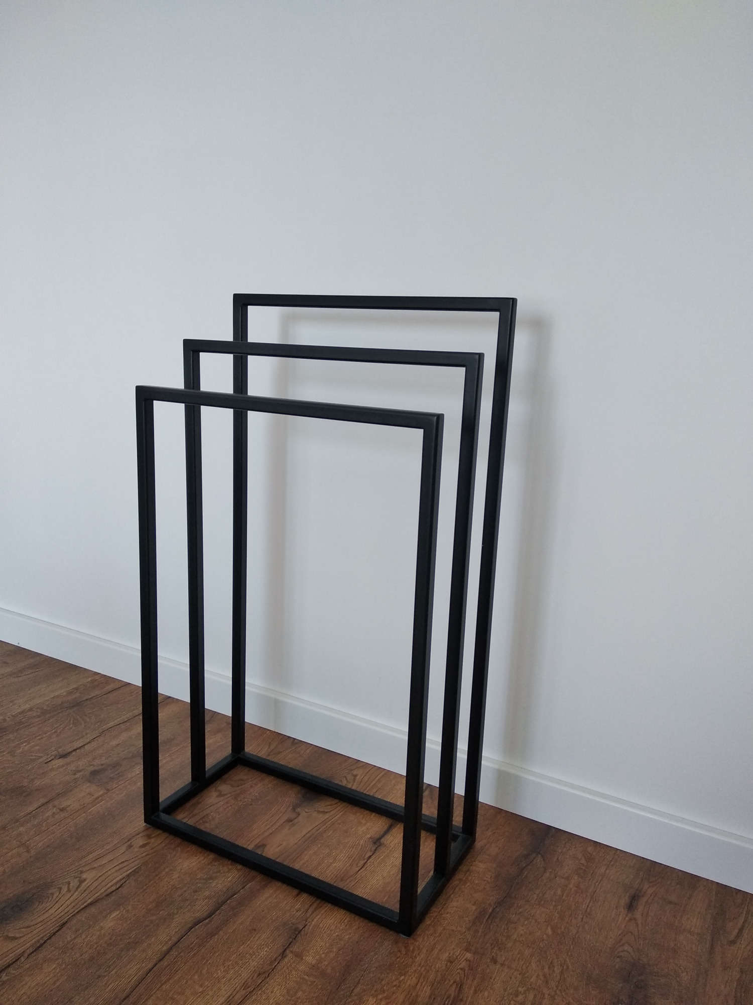 Czarny, metalowy stojak na ręczniki w kształcie trzech prostokątnych ram, ustawiony na drewnianej podłodze na tle białej ściany z listwą przypodłogową.