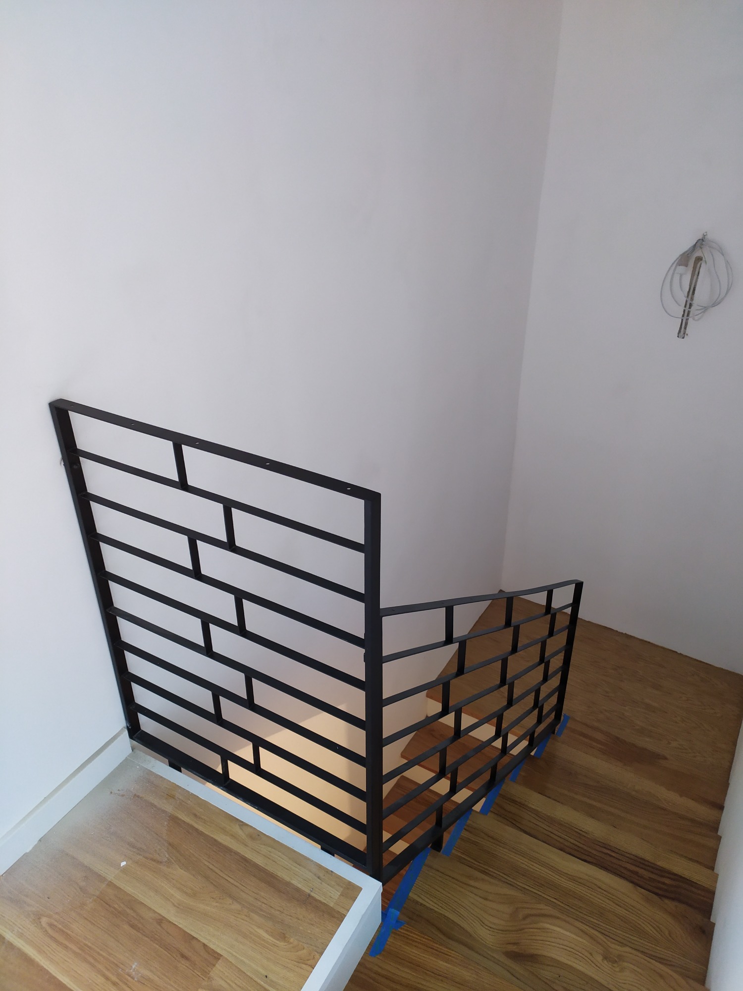 Czarna, geometryczna barierka zabezpieczająca schody z drewnianymi stopniami, widok z góry, niebieska taśma zabezpieczająca na stopniach, surowe wykończenie ścian.