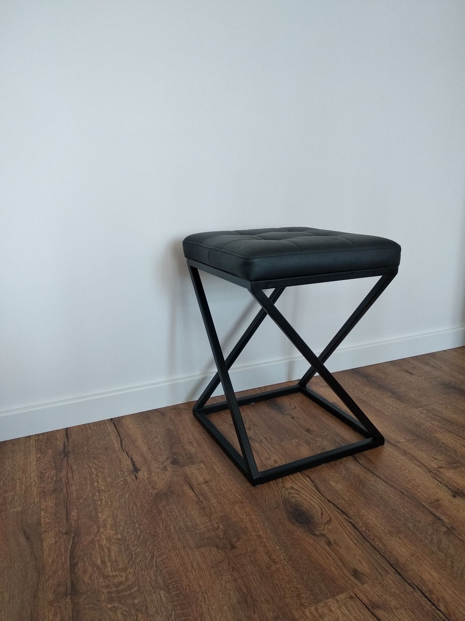 Czarny, tapicerowany taboret z metalową podstawą w kształcie litery X, na drewnianej podłodze przy białej ścianie.