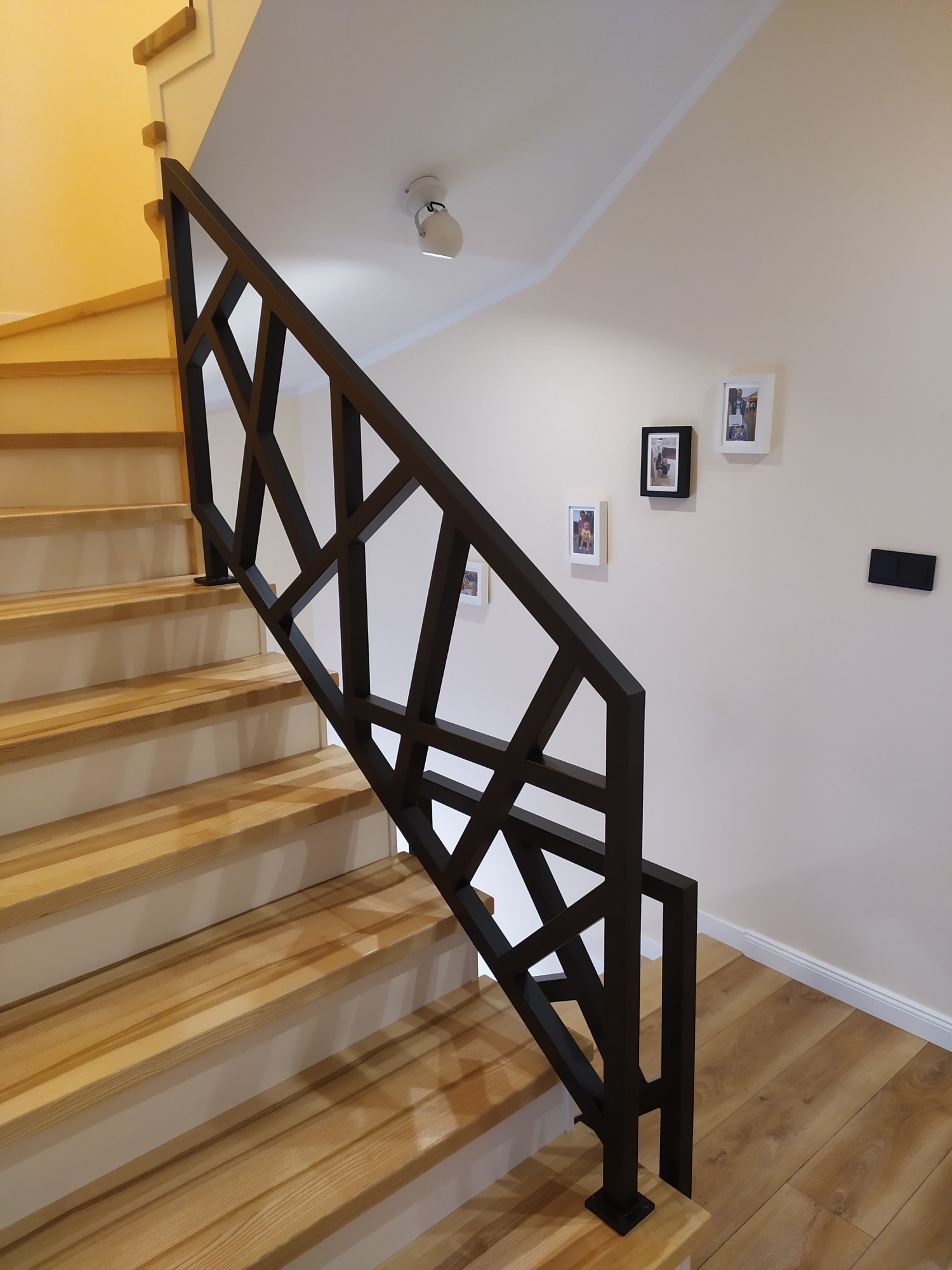 Nowoczesna, czarna konstrukcja zabezpieczająca schody z jasnego drewna, z geometrycznym wzorem, na ścianie trzy małe ramki ze zdjęciami.