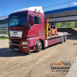 Czerwony ciągnik siodłowy marki MAN z platformą transportową przewożący pomarańczowy w&oacute;zek widłowy, pod wiaduktem, z logo firmy transportowej w lewym g&oacute;rnym rogu kabiny.