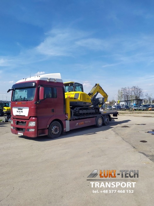 Czerwony ciągnik siodłowy MAN przewozi żółtą koparkę na niskopodwoziowej platformie transportowej firmy ŁUKT-TECH na tle błękitnego nieba.