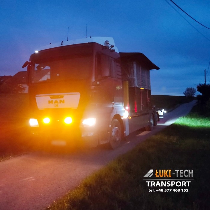 Ciężarówka MAN z włączonymi światłami mijania, przewożąca ładunek zabezpieczony plandeką, na wąskiej asfaltowej drodze o zmierzchu.