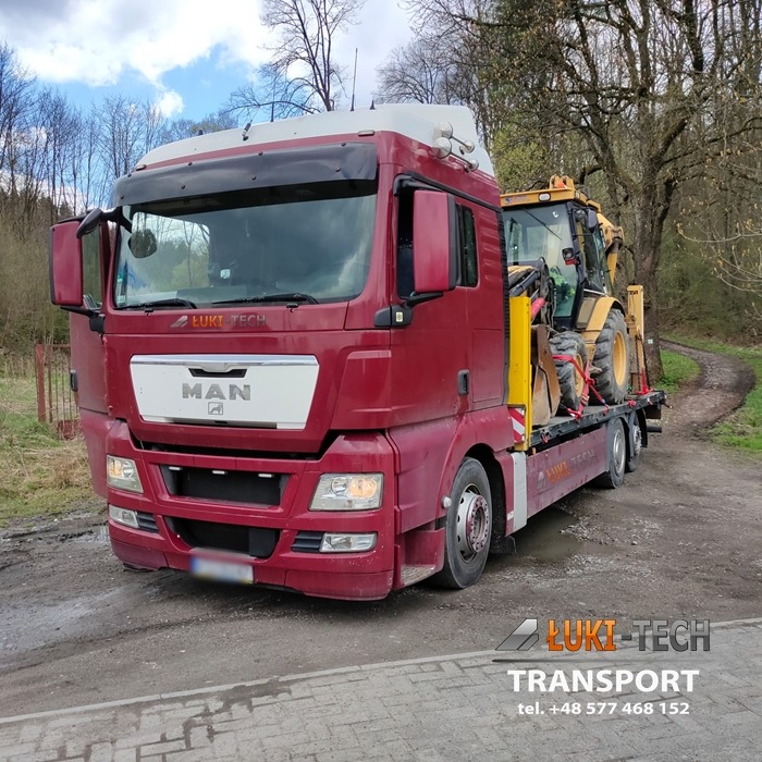 Czerwony ciągnik siodłowy MAN z otwartymi drzwiami kierowcy przewozi na niskopodwoziowej platformie żółtą koparko-ładowarkę, widok z przodu na lewo, w tle drzewa.